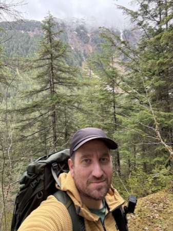 Jeff Zemerick on a forested trail in a yellow fleece and gray cap, carrying a large pack, with a steep conifer covered mountainside and low cloud behind him.
