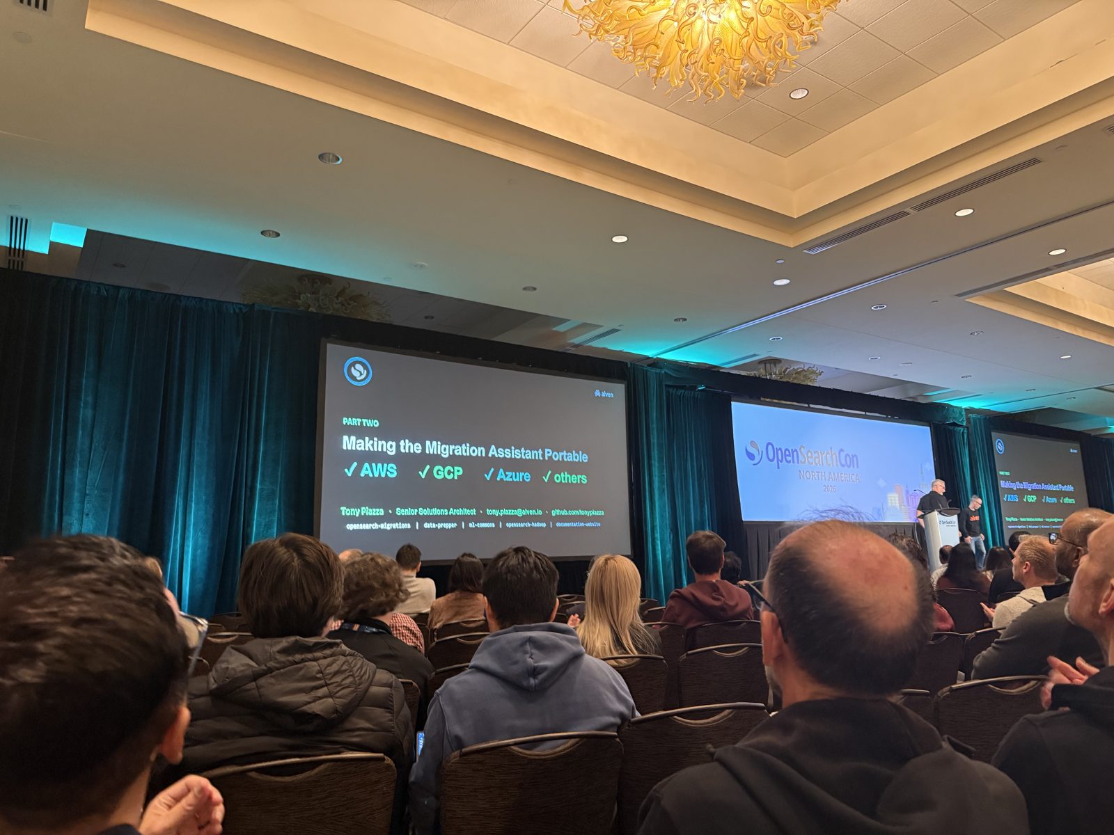 A packed conference ballroom at OpenSearchCon North America 2026. On the main screen, a slide titled Part Two, Making the Migration Assistant Portable, lists AWS, GCP, Azure, and others with check marks above the speaker credit for Tony Piazza of Aiven. An OpenSearchCon North America 2026 logo is on the center screen and presenters stand at the podium.